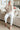 Woman sitting on a couch wearing a beige sweater and white pants in a living room.