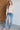 Woman wearing a beige sweater and blue jeans standing against a white brick wall.