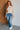 Woman wearing a beige sweater and blue jeans standing against a white brick wall.