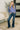 A woman modeling a dusty purple long-sleeve mock neck sweater and dark blue flare jeans, standing in a modern living room setting with a patterned rug, bench, and plant.