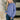 A woman with long blonde hair wearing a dusty purple crewneck sweatshirt and dark blue jeans, standing in a modern indoor setting with a bench and plant in the background.
