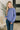 A woman with long blonde hair wearing a dusty purple crewneck sweatshirt and dark blue jeans, standing in a modern indoor setting with a bench and plant in the background.