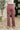 A person shown from the back wearing dusty rose wide-leg corduroy pants, a white top, and white sneakers in an indoor setting with a patterned rug, potted plant, and dark door in the background.