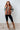 Woman wearing a patterned sweater, brown vest, black leggings, and brown boots against a white wall.