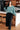 Woman standing in a kitchen wearing a teal sweater and black pants.