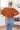 Person wearing a rust-colored sweater and blue jeans in a room with a couch and pillow.