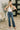 A woman modeling a pair of high-waisted, wide-leg blue jeans with a white sleeveless top, standing in a modern living room setting with a patterned rug and a potted plant.