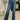 A person wearing medium-wash blue flare jeans with a white top and pink shoes, standing in a bright indoor room with a patterned rug, bench, and potted plant.