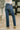 A person shown from the back wearing dark blue straight-leg jeans, a white top, and beige block-heel sandals in an indoor setting with a patterned rug and a potted plant.