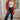 A woman stands indoors wearing a bright orange cardigan with large white floral motifs over a white tank top, blue jeans, and black boots. The photo is taken in a bright room with a window, wood floor, and rug.