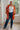 A woman stands indoors wearing a bright orange cardigan with large white floral motifs over a white tank top, blue jeans, and black boots. The photo is taken in a bright room with a window, wood floor, and rug.