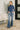 Woman wearing a blue floral top and jeans standing in a room with a white wall and decorative plant.