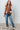 Woman wearing a brown vest, white shirt, blue jeans, and beige boots against a light-colored wall.