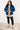 Woman wearing a blue denim jacket, plaid shirt, black pants, and beige boots in a room with white walls.