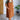 A woman stands indoors modeling a rust-brown quilted pinafore dress with large front patch pockets over a sage green short-sleeve top. She is wearing brown ankle boots, with a white brick wall, wood flooring, a wooden door, and a potted plant in the background.