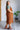 Woman wearing a brown quilted dress and boots in a room with a white brick wall and plant.