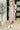 A woman models a dusty rose knit mock-neck midi dress with long draped sleeves, styled with a white belt and white ankle boots in a modern indoor setting.