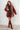 Woman wearing a rust-colored cardigan with fringe, striped dress, and brown knee-high boots against a white paneled wall.