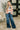 A woman stands indoors wearing a long-sleeve plaid top in peach, gray, and white with wide-leg blue jeans, standing on a patterned rug in front of a black door.