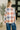 A woman viewed from behind wearing a long-sleeve plaid top with blue jeans, standing indoors near a plant and a dark door.