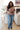 Woman standing in a living room wearing a brown sweater and blue jeans.
