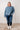 Woman wearing a blue cable knit sweater and jeans standing against a white paneled wall.