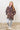 Person wearing a leopard print hoodie and light gray pants against a white wall.