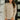 A woman wearing a beige cable-knit crewneck sweater and mauve leggings, standing in a modern indoor setting with white walls and black-framed windows.