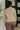 Person wearing a beige sweater and brown pants standing in a room with a plant and black screen in the background.