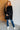 Woman wearing a black oversized coat against a white brick wall.