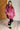 Woman wearing a pink oversized hoodie in a room with wooden floor and white brick wall.
