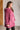 Woman wearing a pink hooded garment against a white brick wall.