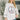 A woman wearing a white crewneck sweatshirt with a large peace sign graphic on the front made of red, blue, and orange flowers, paired with blue jeans in a bright indoor setting.
