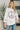 A woman wearing a white crewneck sweatshirt with a large peace sign graphic on the front made of red, blue, and orange flowers, paired with blue jeans in a bright indoor setting.