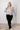 Woman wearing a patterned blouse and black pants against a white wall.