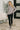 Woman wearing a gray sweater and black leggings standing in a room with a plant and bench.