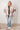 Person wearing a patterned jacket and jeans standing against a white tiled wall.