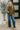 Woman wearing an olive green blouse and blue jeans standing next to a potted plant indoors.