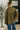 Person wearing an olive green jacket and blue jeans standing in a room with a plant and framed picture on the wall.