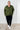 Person wearing a green hoodie and black pants against a white paneled wall.