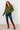Woman wearing an olive green hoodie, blue jeans, and brown knee-high boots against a white brick wall.