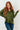 A woman with long wavy red hair wearing an olive green long-sleeve polo-style top and blue jeans, standing against a white paneled wall.
