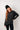 Woman wearing a dark gray sweater and black beanie against a white tiled wall.