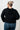 Person wearing a black textured hoodie and blue jeans against a white tiled wall.