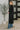 Woman wearing a long black coat and blue jeans standing next to a potted plant indoors.