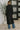 Person wearing a black coat and blue jeans standing in a room with a plant and abstract wall art.