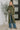 A woman modeling a long-sleeve, high-neck, olive green knit cowl-neck sweater with a front slit, styled with wide-leg blue jeans and brown shoes in a modern indoor setting.