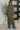 Person wearing a long green cardigan over blue pants in a room with a plant and abstract wall art.