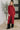 A woman models a long-sleeve, cowl-neck, midi-length knit sweater in heathered red. The sweater features a draped wrap-style front panel over a sheer, dark lining.