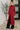 Woman wearing a long red dress standing indoors with a plant in the background